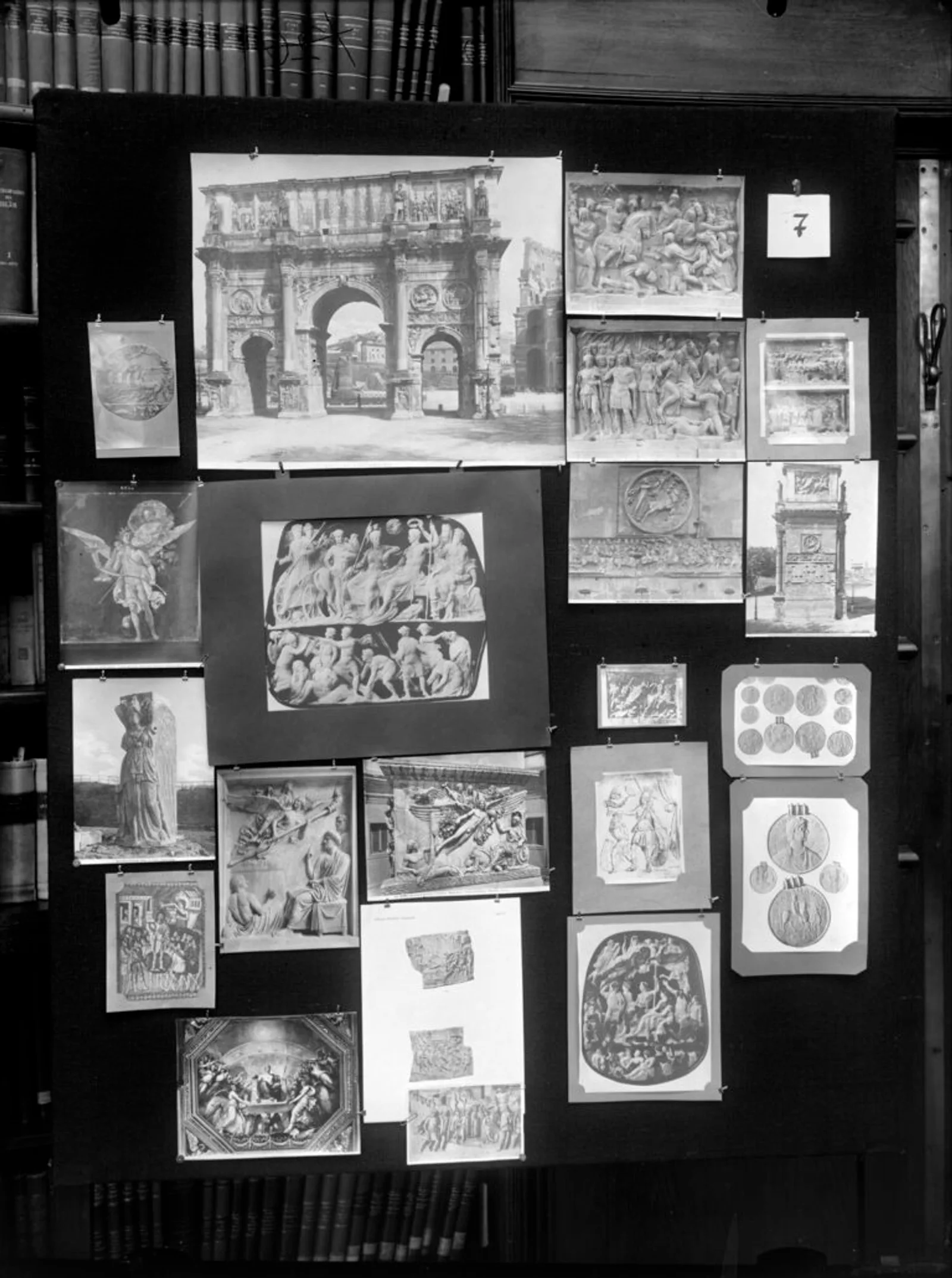 Photograph of panel 8 of Aby Warburg's Mnemosyne Atlas: a large black cloth board mounted in front of library bookshelves, pinned with roughly twenty black-and-white photographic reproductions of varying sizes, mostly Roman triumphal reliefs, sarcophagi.